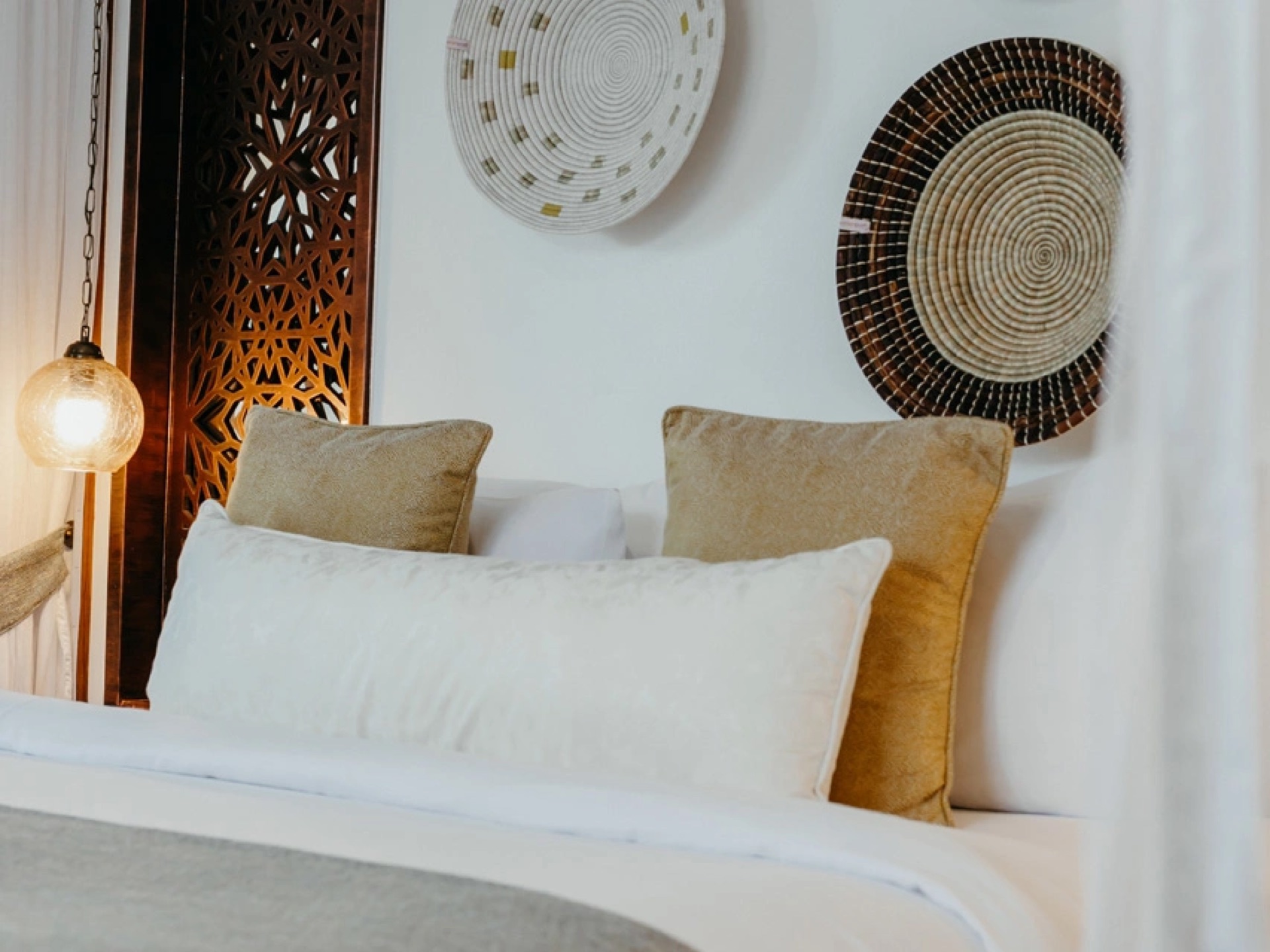 A bed with a carved wooden screen headboard, woven wall baskets and a pendant lamp in a Zanzibar hotel room