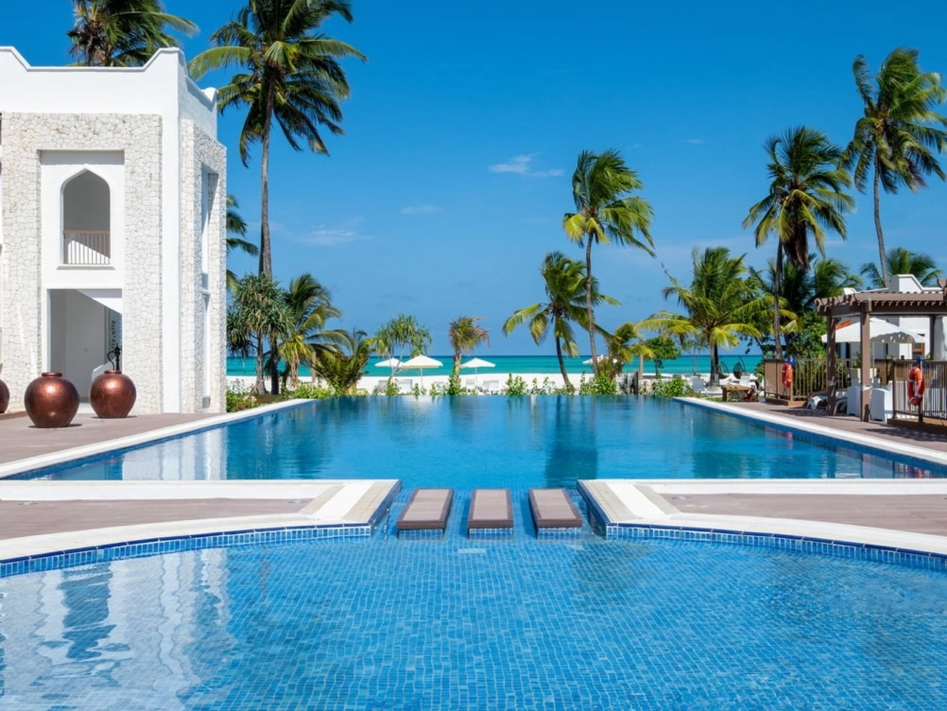 A long swimming pool between whitewashed Swahili-arched buildings, with coconut palms and white sand beyond