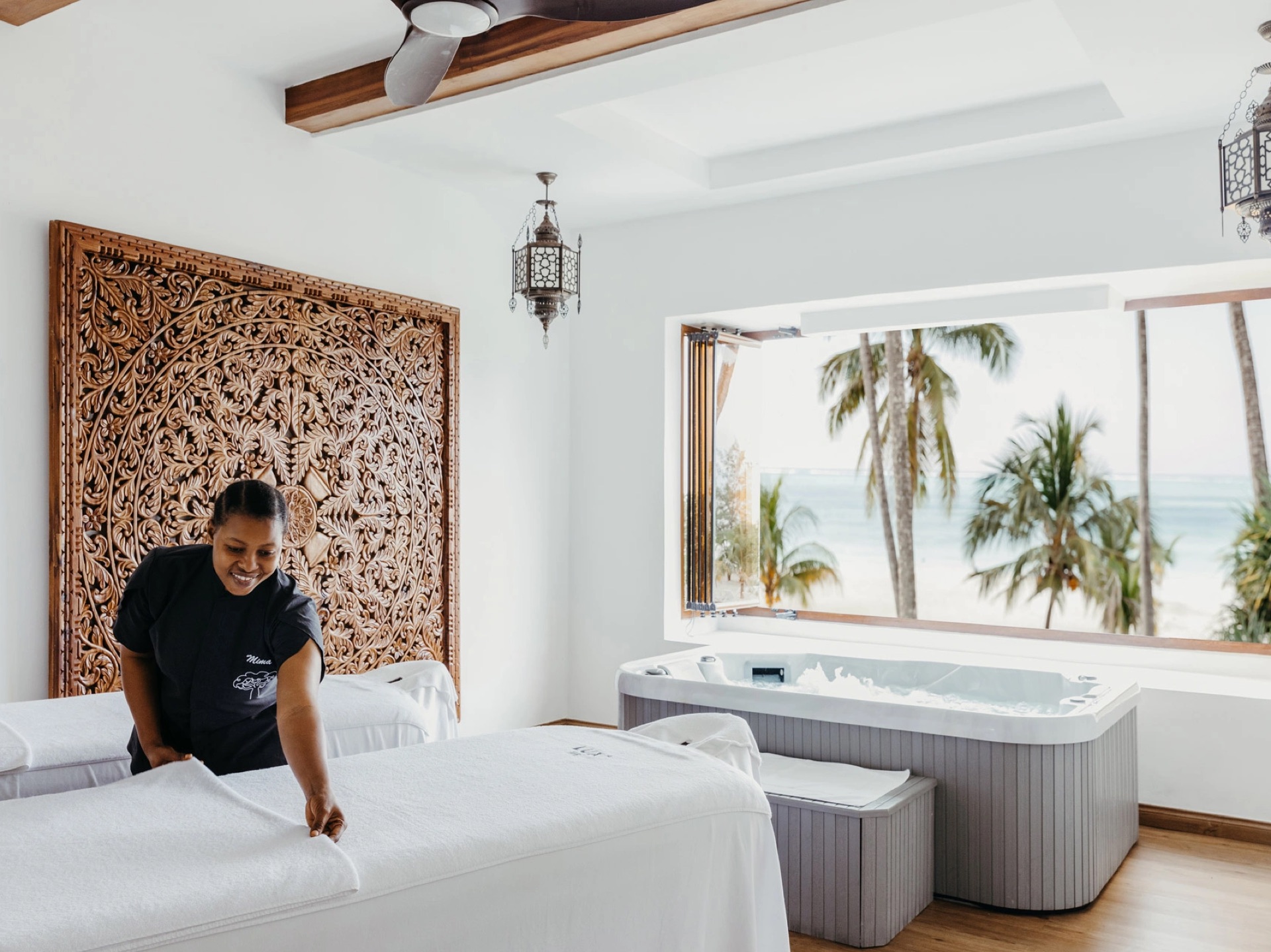 A therapist preparing twin massage beds in a spa room with a carved wooden screen and an open window onto the sea